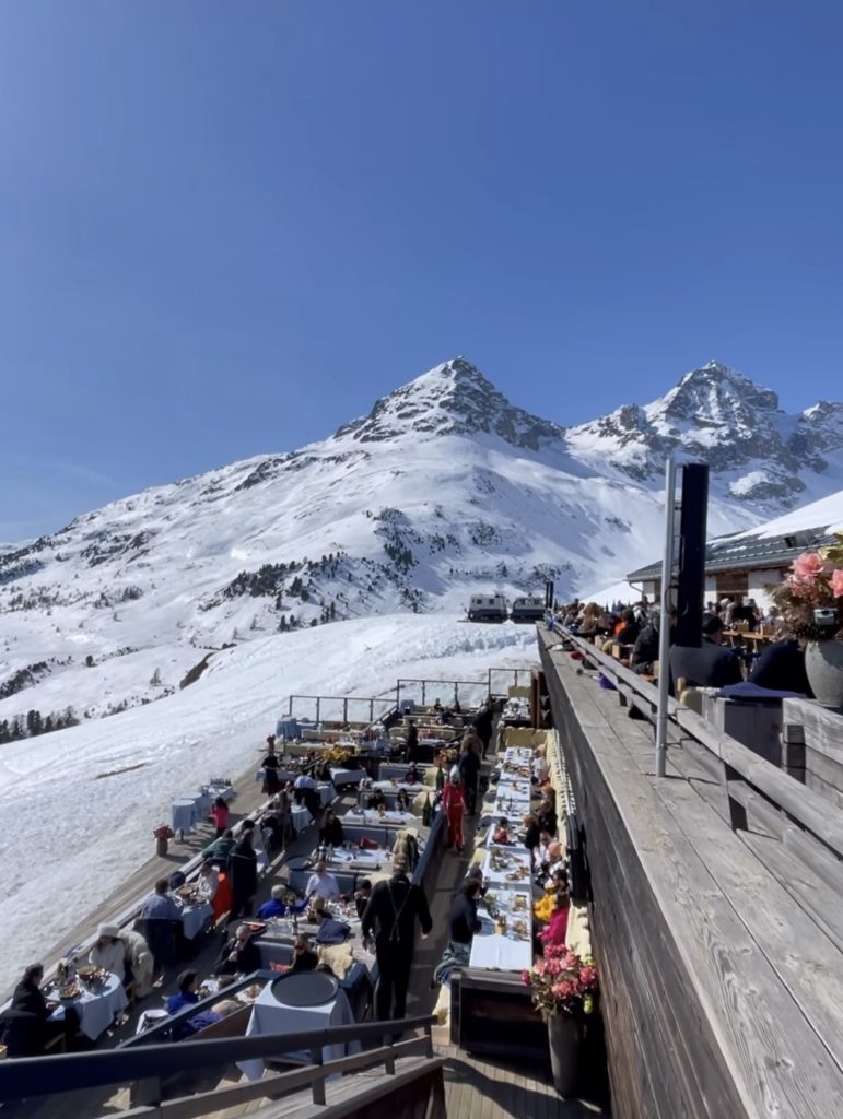 Busy mountain restaurant terrace in St Moritz with guests dining outdoors in winter, overlooking snowy Alpine peaks