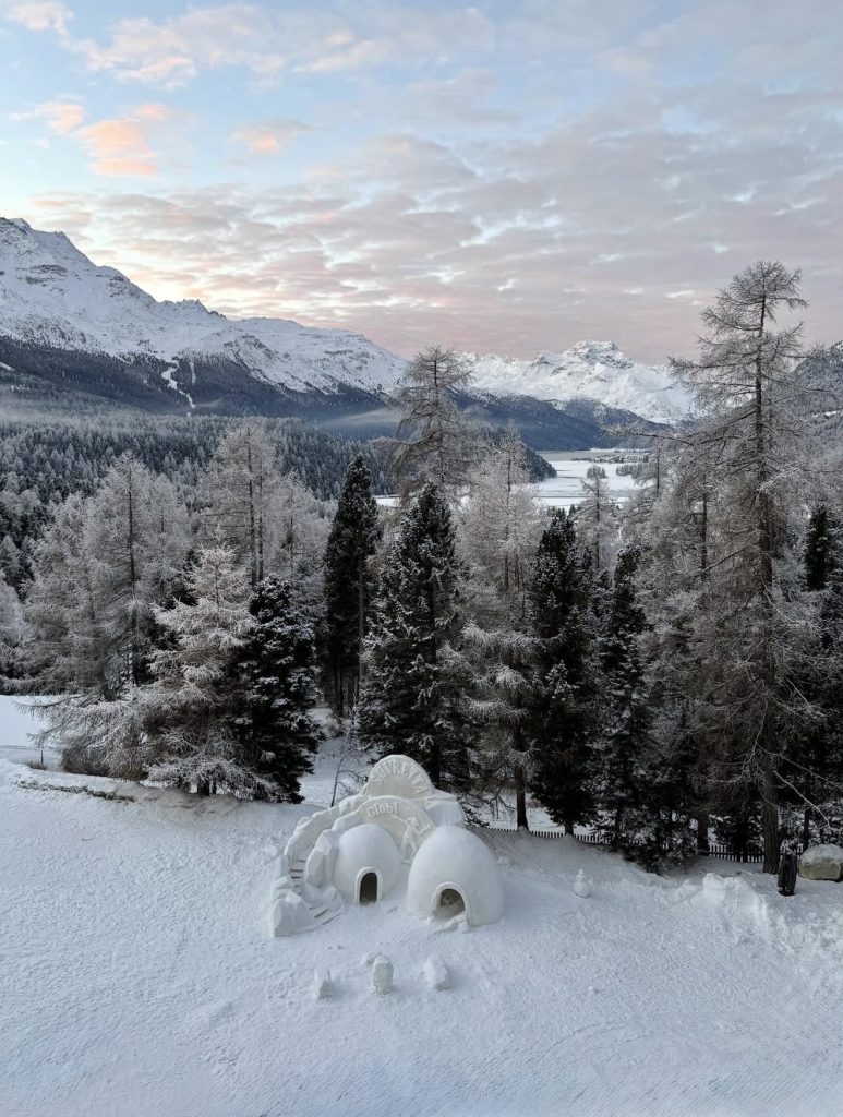 Winter landscape in St Moritz with snowy forest, mountains, and an igloo structure in the foreground at sunrise.