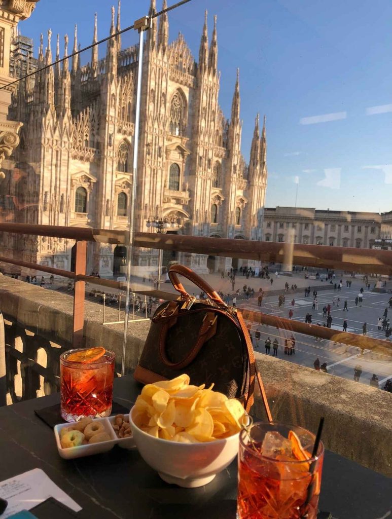 a picture of a rooftop bar with snacks and drinks on the table with the Duomo backdrop